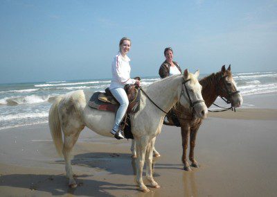 Two girls on horseback at the beach with waves in the background.