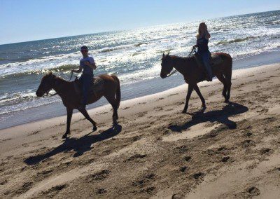 Two individuals riding horses along a sunny beach shoreline.